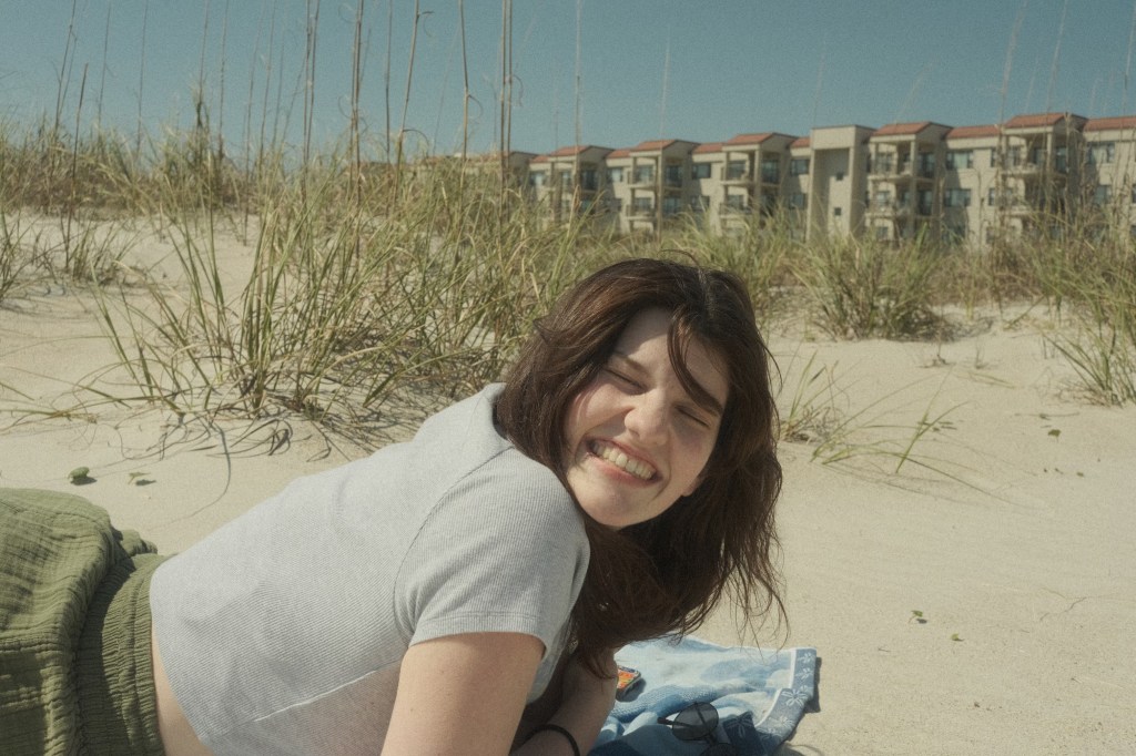 A photo of a girl smiling with her eyes closed on a sand dune, wearing a grey tee and green pants.