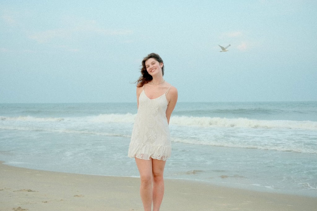 a photo of tall girl with brown hair in front of the ocean with a seagull flying in the air